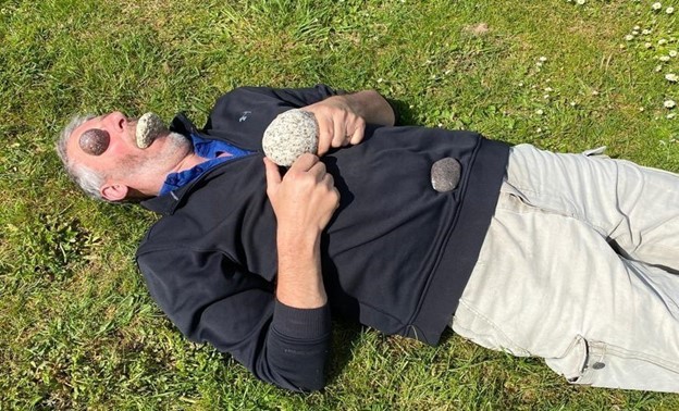 man lying on grass with stones on eyes, in mouth, on chest and stomach